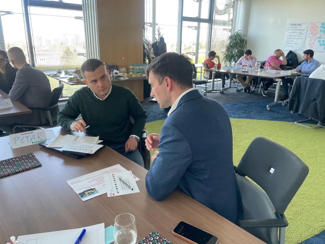 Geschäftstreffen und Besprechung im Team Zwei Männer unterhalten sich während eines Arbeitstreffens am Tisch, während im Hintergrund andere Teilnehmer in Gruppen arbeiten